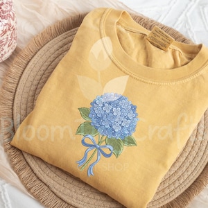 May include: Mustard yellow sweatshirt featuring a blue hydrangea bouquet with green leaves and a blue ribbon. The floral design is centered on the front of the shirt. The shirt is folded on a woven tray.