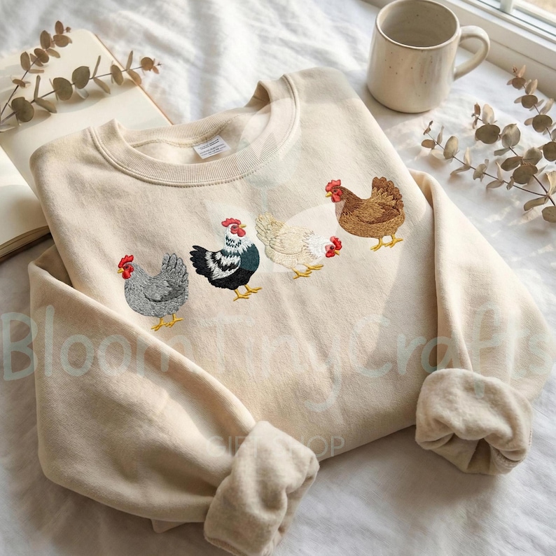 May include: Cream-colored sweatshirt featuring an embroidered design of four chickens in various colors. The chickens are in shades of gray, black and white, white, and brown, each with a red comb. The sweatshirt has a crew neck and long sleeves.