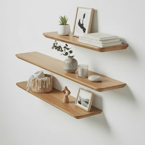 Puede incluir: Tres estantes flotantes de madera montados en una pared blanca, cada uno con objetos decorativos. Los estantes exhiben una pequeña planta en maceta, arte enmarcado, libros, un jarrón, una vela, una cesta tejida y una escultura de madera.