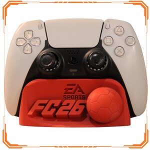Può includere: Un controller di gioco bianco e nero è appoggiato su una base rossa con la scritta "EA SPORTS FC26" e un pallone da calcio. Il controller ha pulsanti e pulsanti direzionali. La base ha il logo EA Sports.