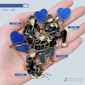 May include: Enamel pins featuring stylized figures in black jackets, holding cameras, with blue glitter heart accents. Text includes "VOCAL LINE", "PLAYER" names, and "PEW PEW!". Gold chain detail.