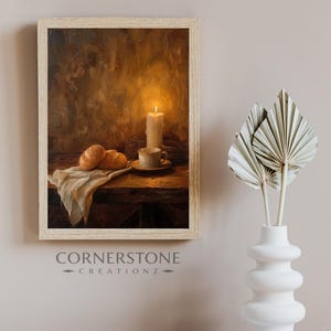 Puede incluir: Pintura enmarcada de naturaleza muerta que representa una vela encendida, una taza y un platillo, y dos cruasanes sobre una mesa de madera. Un paño blanco está sobre la mesa. La pintura tiene una paleta de colores cálidos.