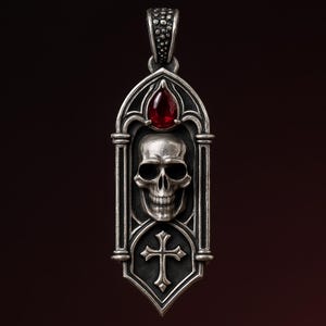 Puede incluir: Colgante plateado con diseño de calavera. El colgante tiene forma de arco gótico, una gema roja y una cruz en la parte inferior. La calavera está detallada con dientes y cuencas de los ojos visibles. El colgante cuelga de una fianza texturizada.