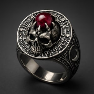 May include: A silver skull ring with a red gemstone. The ring features a detailed skull design with intricate carvings and a circular band with engraved symbols. The gemstone is held by skeletal hands.