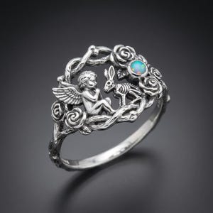 Puede incluir: Un anillo de plata con un querubín alado frente a un conejo esquelético, rodeado por un marco de rosas y enredaderas retorcidas. Un pequeño ópalo redondo e iridiscente está engastado en el diseño floral, añadiendo un toque de color al metal.