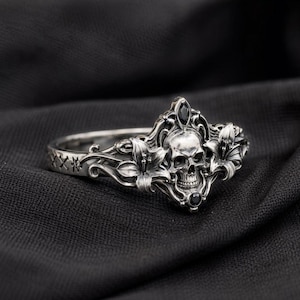 May include: A silver ring with a skull design and black gemstone embellishments. The ring features detailed floral and vine patterns, a skull centrepiece, and black gems. The ring is set against a dark backdrop.