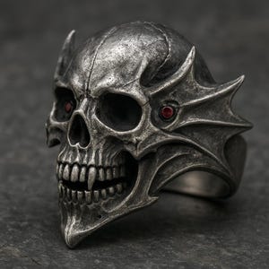 Könnte beinhalten: Ein silberner Totenkopfring mit roten Edelsteinaugen und dekorativen Seitenakzenten. Der Ring hat ein detailliertes Totenkopfdesign mit Fangzähnen und einer strukturierten Oberfläche.