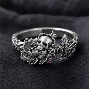 May include: A silver ring featuring a skull with a crown, set within a floral design. The ring has intricate detailing, a small red gem, and a black gem. The ring's design includes a fleur-de-lis element.