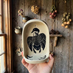 May include: Cream-colored ceramic soap dish with a black loon illustration. The dish has an oval shape with a small hole at the top for hanging. The loon design features detailed wing patterns. The background is a rustic wooden wall.