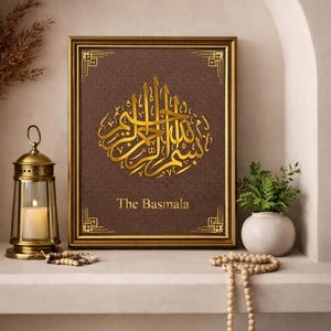 Op de afbeelding: Een ingelijst kunstwerk met gouden kalligrafie op een bruine achtergrond, met de tekst "The Basmala" eronder. De lijst is goudkleurig. Het kunstwerk wordt tentoongesteld op een plank met decoratieve items, waaronder een lantaarn en een kleine plant.