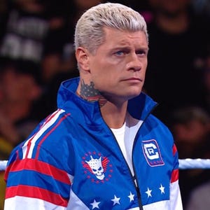 May include: A blue, white, and red jacket with a skull and star emblem. The jacket has a zipper and a high collar. The person in the image has blonde hair and a neck tattoo.
