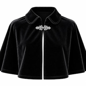 May include: A black velvet capelet featuring a rounded collar and a silver-toned metal filigree clasp at the neckline. The garment has a short, flared silhouette with wide, open sleeves that reach the mid-torso, providing a structured drape.