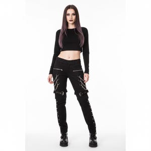 May include: Black pants with silver chain and buckle details. The pants have zippers on the front pockets and decorative chains hanging from the sides. The pants are paired with a black long-sleeve crop top and black platform boots.