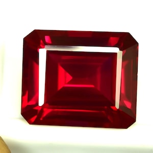 Peut inclure: Une grosse pierre précieuse rubis de coupe rectangulaire, d'un rouge profond. La gemme a une surface à facettes qui reflète la lumière, créant un éclat brillant. Les bords sont coupés avec précision, mettant en valeur la clarté et la couleur de la gemme.