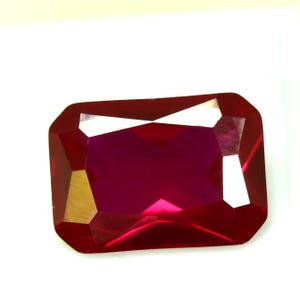 Può includere: Una pietra preziosa di rubino tagliata a forma rettangolare sfaccettata. La gemma è di un colore rosso intenso e saturo con un alto grado di chiarezza. Il taglio presenta una serie di sfaccettature angolate che riflettono la luce.