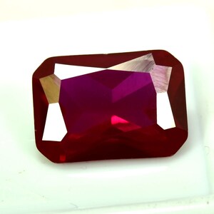 May include: A faceted, rectangular ruby gemstone with a deep red hue. The gem has a polished surface with sharp edges and internal reflections, showcasing its clarity and brilliance. The gemstone is set against a white background.