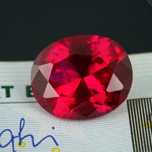 Könnte beinhalten: Ein facettierter, oval geformter, tiefroter Edelstein. Der Edelstein hat mehrere Facetten, die das Licht reflektieren und einen brillanten Glanz erzeugen. Der Edelstein liegt vor einem Hintergrund mit grünem und blauem Text.