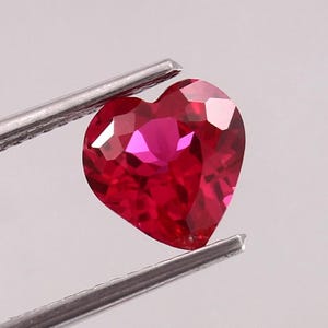 May include: A heart-shaped, faceted ruby gemstone held by tweezers. The ruby is a deep, vibrant red with multiple facets that reflect light. The gemstone is a symbol of love and passion.