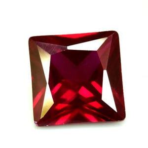 Puede incluir: Una gema tallada cuadrada y facetada, de color rojo intenso. La gema tiene bordes afilados y una superficie pulida, que refleja la luz para crear reflejos y sombras. La gema está sobre un fondo blanco.