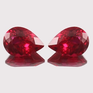 May include: Two faceted, pear-shaped, deep red gemstones are displayed. The gems are highly polished, reflecting light and creating a brilliant sparkle. The stones are set against a light background, with their reflections visible below.