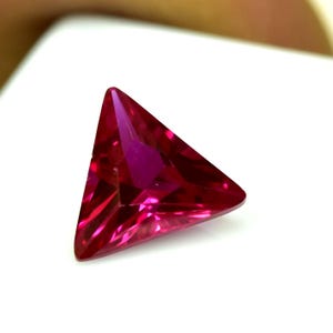 May include: A triangular-cut gemstone in a rich, deep red colour. The facets catch the light, creating a sparkling effect. The gem's shape is a precise equilateral triangle, highlighting the quality of the cut and the stone's clarity.
