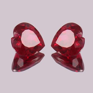 Puede incluir: Dos gemas facetadas en forma de corazón, de un tono rojo intenso. Las gemas se muestran sobre una superficie reflectante, creando un efecto de espejo. Las piedras tienen un aspecto pulido y brillante.
