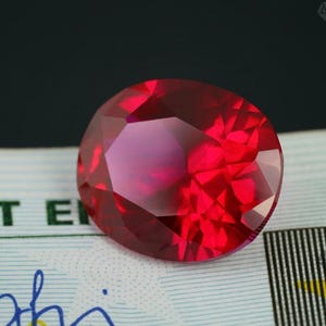 Op de afbeelding: Een gefacetteerde, ovaal geslepen robijn edelsteen. De edelsteen heeft een levendige rode kleur, met meerdere facetten die licht reflecteren. De robijn is geplaatst tegen een achtergrond met patronen, met een donkere achtergrond.