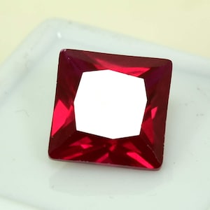 Peut inclure: Une pierre précieuse rouge foncé, taillée en carré et à facettes, avec un centre blanc brillant. La pierre est posée sur un fond blanc, mettant en valeur sa couleur vibrante et sa clarté. Les bords de la gemme sont nets et bien définis.