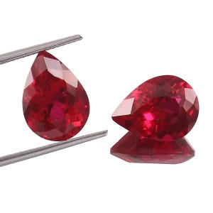 May include: Two pear-shaped, faceted, deep red gemstones. One is held by silver tweezers, the other rests on a reflective surface, showcasing its brilliance. The gems exhibit a rich, saturated color and a high degree of clarity.