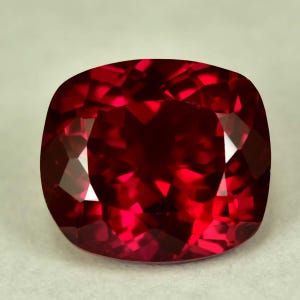 May include: A cushion-cut, faceted gemstone with a deep red hue. The gem's facets reflect light, creating a brilliant display of color and sparkle. The shape is a square with rounded corners, giving it a soft, elegant appearance.