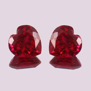 Könnte beinhalten: Zwei herzförmige, facettierte Edelsteine in leuchtendem Rot. Die Edelsteine werden auf einer reflektierenden Oberfläche präsentiert, was ihren Glanz verstärkt. Der Schliff der Steine erzeugt mehrere Facetten, die das Licht reflektieren und ihren Glanz verstärken.