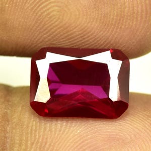 Könnte beinhalten: Ein facettierter, rechteckiger Rubin-Edelstein mit tiefroter Farbe. Der Edelstein hat eine helle, reflektierende Oberfläche mit scharfen Kanten und einem polierten Finish. Der Rubin wird zwischen zwei Fingern gehalten.