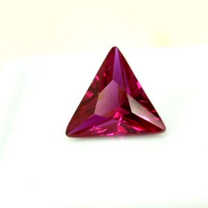 May include: A triangular, faceted gemstone in a deep magenta colour. The gem is cut with multiple facets, creating a brilliant display of light and colour. The gemstone is set against a white background.