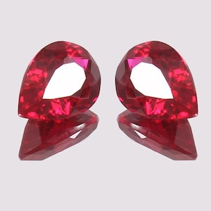 May include: Two faceted, pear-shaped rubies in a deep red colour. The gems are displayed on a reflective surface, highlighting their cut and brilliance. The rubies have a large, white, teardrop-shaped facet in the centre.