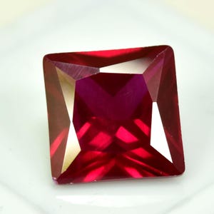 Pode incluir: Uma pedra preciosa facetada de corte quadrado, com um tom vermelho profundo. A gema tem bordas afiadas e uma superfície polida, refletindo a luz para criar uma exibição brilhante. A pedra preciosa está sobre um fundo branco.