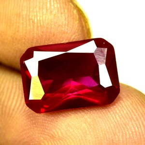 Pode incluir: Uma pedra preciosa de rubi retangular e facetada. A gema é de cor vermelha profunda com reflexos brilhantes e uma superfície polida. O corte apresenta múltiplas facetas que refletem a luz, criando uma aparência brilhante. A pedra preciosa é segurada pelos dedos de uma pessoa.