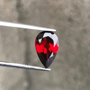 Può includere: Una gemma sfaccettata a forma di pera tenuta da una pinzetta. La gemma mostra un colore rosso intenso con aree scure, quasi nere. Il taglio della pietra crea molteplici sfaccettature che riflettono la luce, esaltando la sua brillantezza. Lo sfondo è grigio neutro.