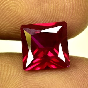 Puede incluir: Una piedra preciosa de rubí tallada en forma cuadrada y facetada. La gema es de un rojo intenso y vibrante con facetas nítidas y reflectantes. La piedra preciosa se sostiene entre los dedos, mostrando su brillo y claridad. Adecuado para la fabricación de joyas.