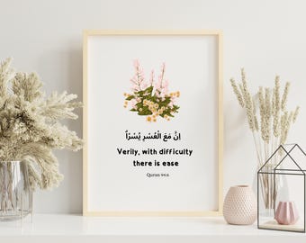 May include: A framed print with a light wood frame featuring floral artwork and Arabic calligraphy with English translation. The text reads "Verily, with difficulty there is ease" from Quran 94:6. The artwork includes pink and yellow flowers.