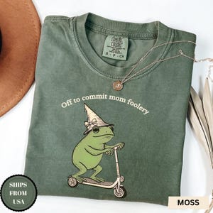May include: Moss green t-shirt featuring the text "Off to commit mom foolery" and a cartoon frog on a scooter, wearing a witch's hat. A silver necklace is draped across the shirt.