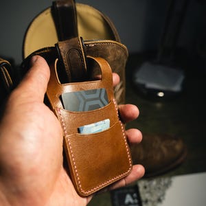May include: A tan leather card holder featuring two vertical slots, light pink stitching, and a top cutout. The holder contains a dark grey card with a geometric pattern and a folded light blue paper bill tucked into the front pocket.
