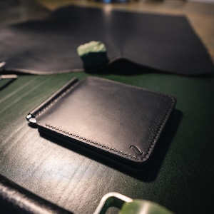 May include: A black leather money clip wallet with a silver clip, featuring a stitched border and a small embossed design. The wallet is on a green surface, with leather pieces and tools in the background.