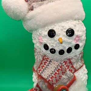 May include: A whimsical snowman figurine with a red Santa hat and a patterned scarf. The snowman has black button eyes and a carrot nose. The scarf has red, white, and gray patterns. The snowman is a festive holiday decoration.
