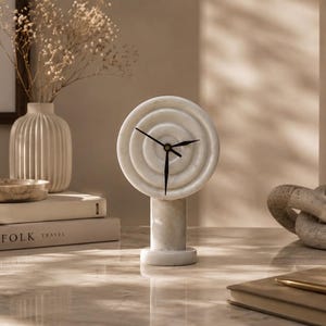 May include: A white marble table clock featuring a circular face with concentric ring carvings and a cylindrical base. The clock has three thin black hands positioned at the centre of the circular face, which is mounted atop a solid marble pedestal.