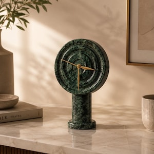 Può includere: Un orologio circolare in marmo verde caratterizzato da un motivo testurizzato a cerchi concentrici sul quadrante. L'orologio è sostenuto da una base cilindrica in marmo e presenta tre sottili lancette in metallo dorato al centro.