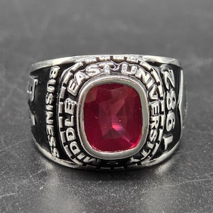 May include: A silver-toned ring with a large, square, ruby-red gemstone at its center. The ring is engraved with the words "MIDDLE EAST UNIVERSITY BUSINESS" and the year "1986".