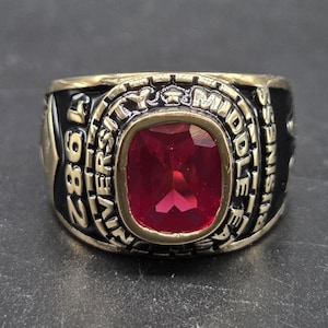 May include: A gold-toned ring with a large, rectangular, red gemstone at the center. The ring has black enamel on the sides with the words "UNIVERSITY MIDDLE EAST" and "BUSINESS" inscribed. The year "1982" is also visible.