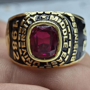 May include: A gold-toned ring with a large, rectangular, deep red gemstone at its center. The ring has black enamel sides with the text "MIDDLE EAST BUSINESS UNIVERSITY 1982" in gold lettering. A classic signet ring.
