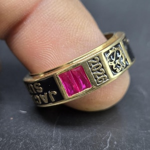 May include: A gold-colored ring with a rectangular, magenta-colored stone. The ring has black enamel sections with the text "JAC SU" and "2026". Decorative patterns are etched into the gold band.