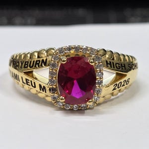 May include: A gold-colored class ring with a large, oval, ruby-red stone at the center, surrounded by a halo of smaller clear stones. The ring has the words "RAYBURN" and "HIGH SCH" engraved on it, along with "MI LEU M" and the year "2026".